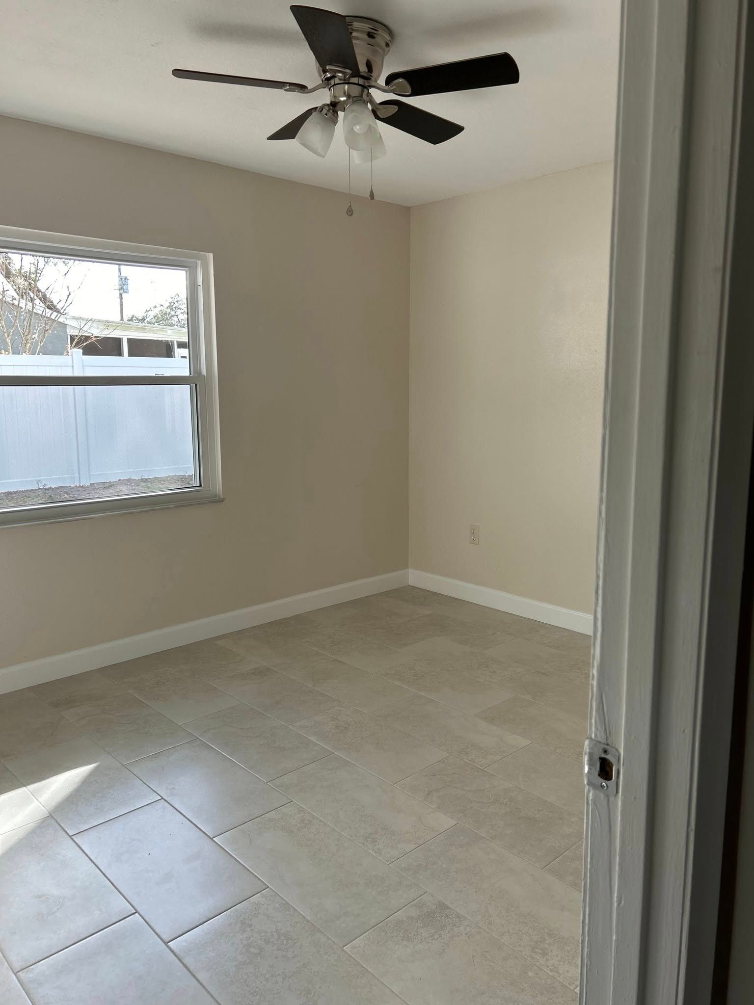 Empty bedroom with tiled floor, window, ceiling fan, and off-white walls.