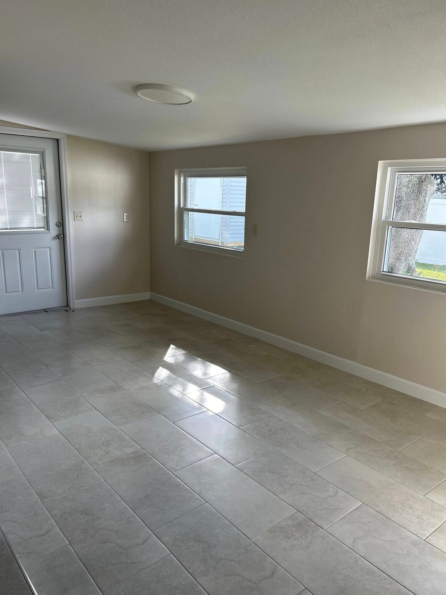 An empty room with light-colored walls, gray tiled floor, two windows, and a white door.