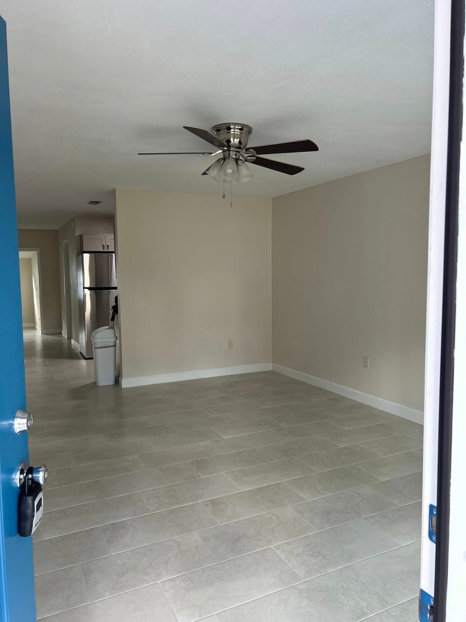 View from a blue doorway into a mostly empty room with a ceiling fan; a kitchen is in the background.