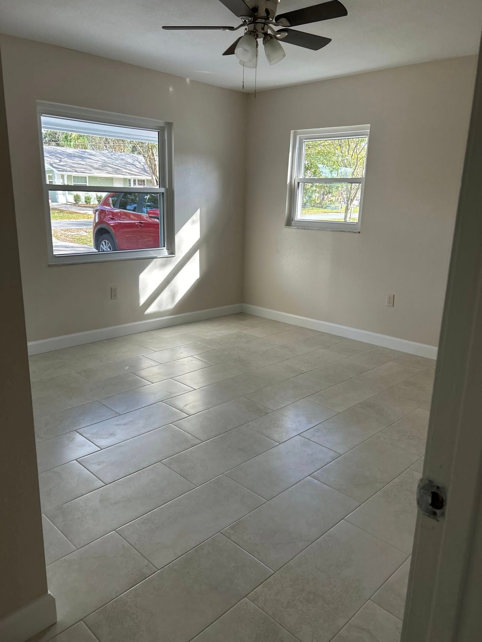 Empty bedroom with tile floor, two windows, ceiling fan, and beige walls.