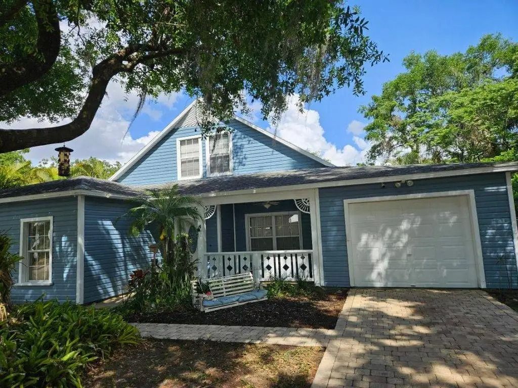 Blue house with white trim, porch, garage, and walkway under a blue sky with trees.