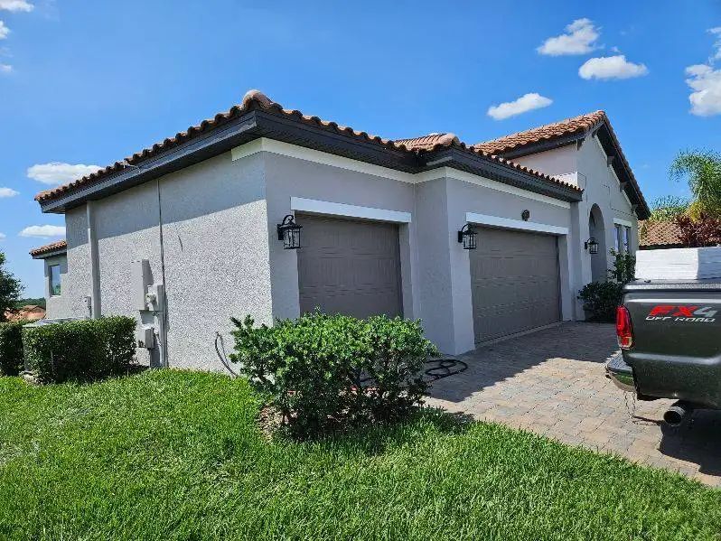 A light gray stucco house with a brown tile roof and garage doors under a blue sky.