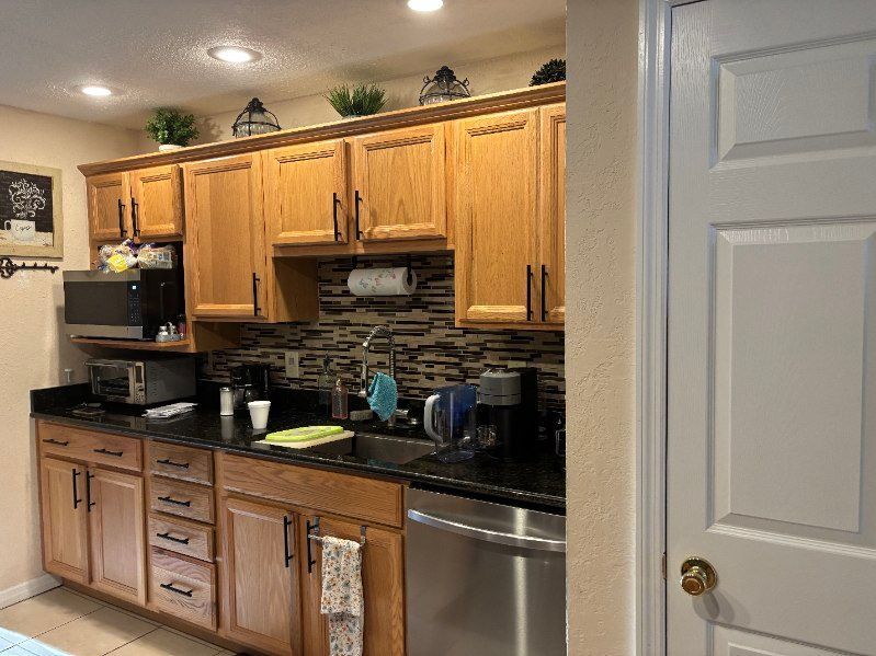 A kitchen with light brown cabinets, black countertops, and a stainless steel dishwasher, with a white door on the right.