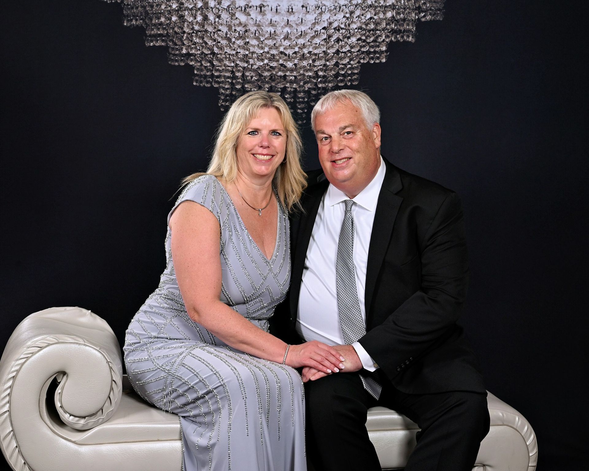 Couple seated on a white chaise lounge, smiling, formal attire, under a chandelier.