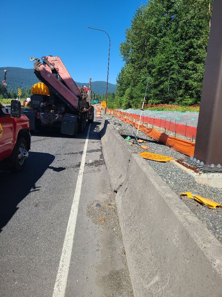 Roadside scene: crane truck, concrete barrier, orange construction barriers, and green trees.