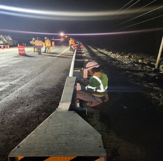 Road workers in reflective vests install a barrier on a highway at night, illuminated by overhead lights.