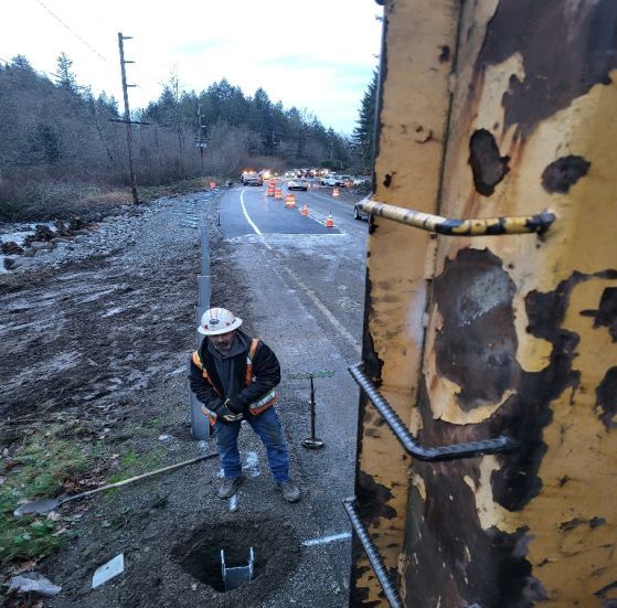 Construction worker installing a utility pole next to a road, with orange cones and other vehicles visible in the distance.