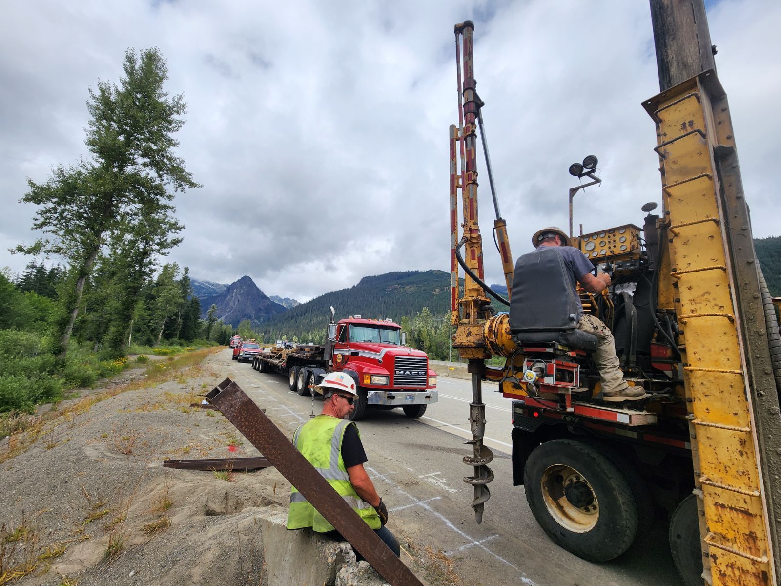 Workers drilling holes on a road. One operates a yellow drill, another watches. A red truck and mountains are in the background.