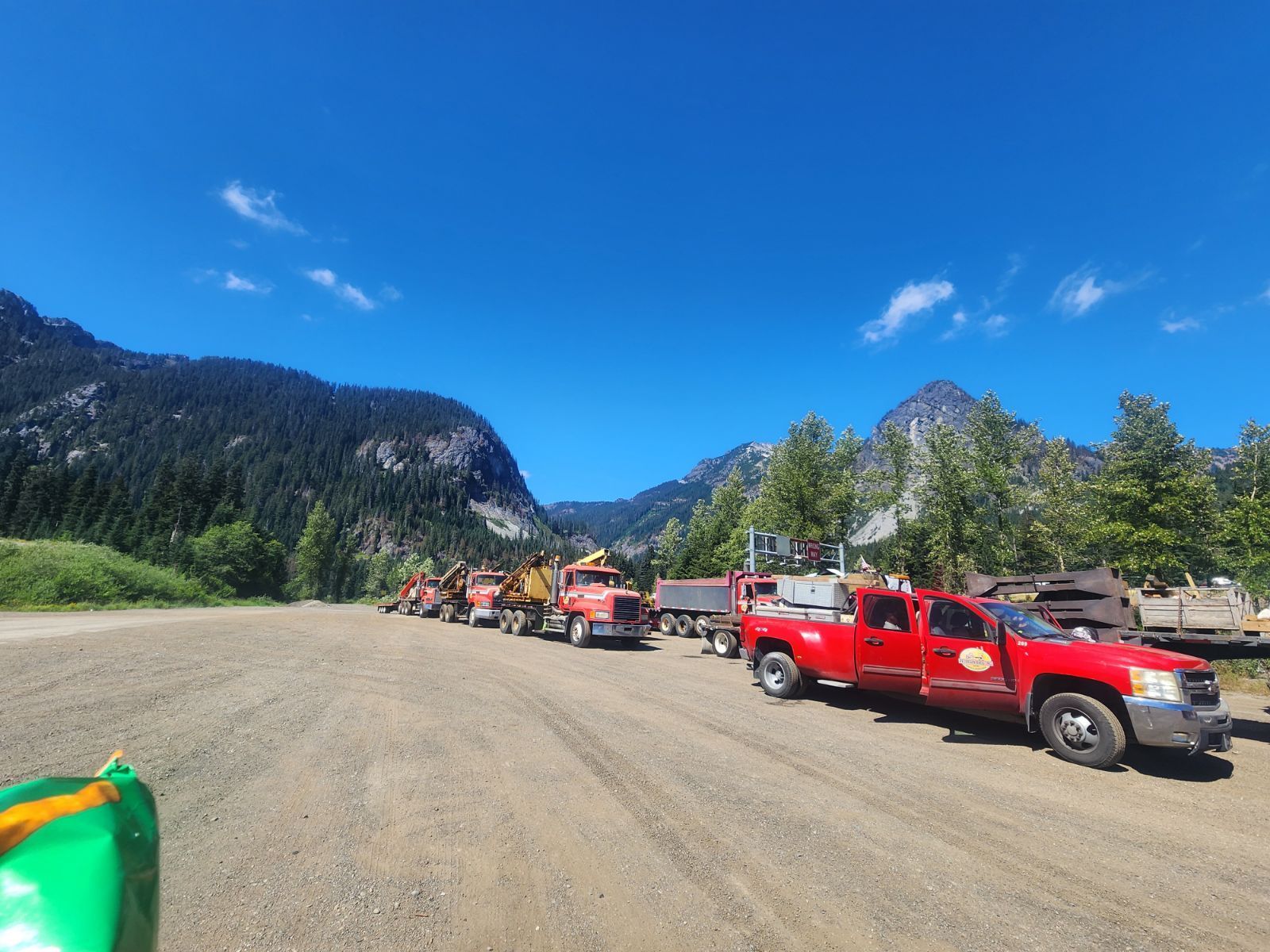 Red trucks and machinery parked on a gravel road against a backdrop of mountains and a blue sky.