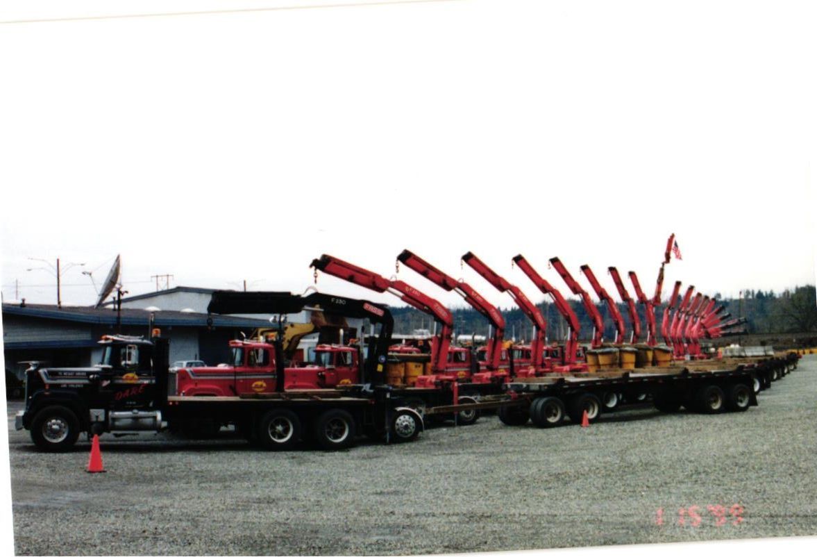 Line of red and black trucks with cranes parked on gravel.