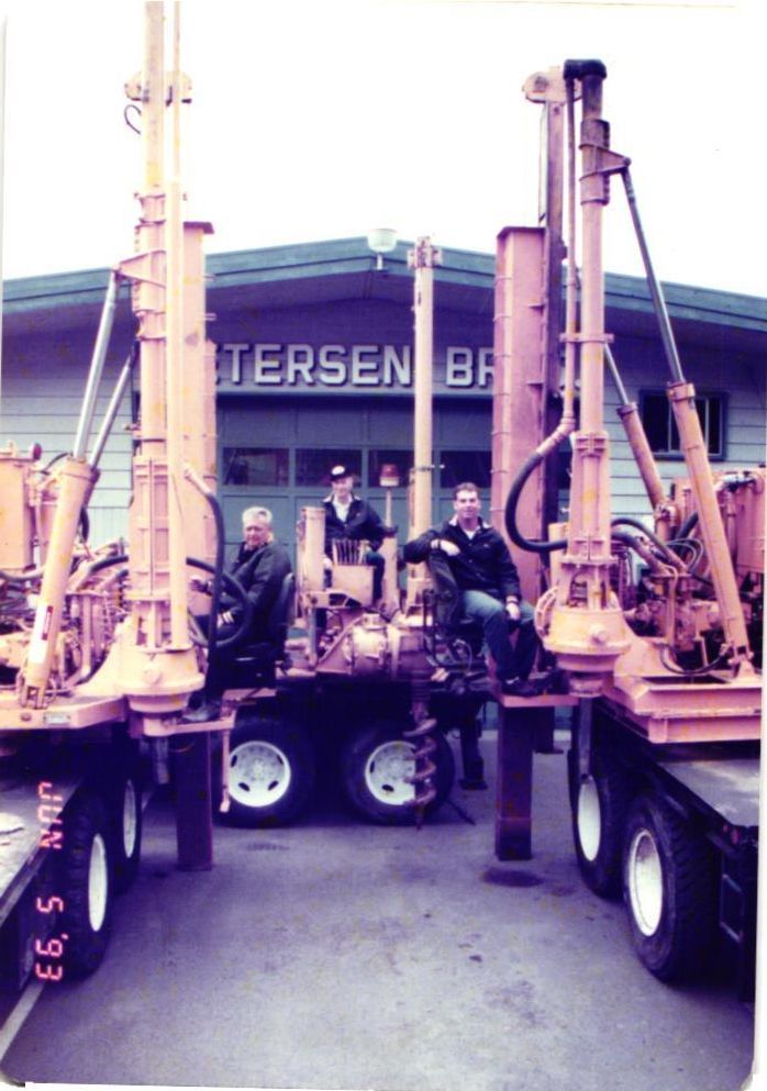 Three men pose near two orange drilling rigs in front of a building with the name