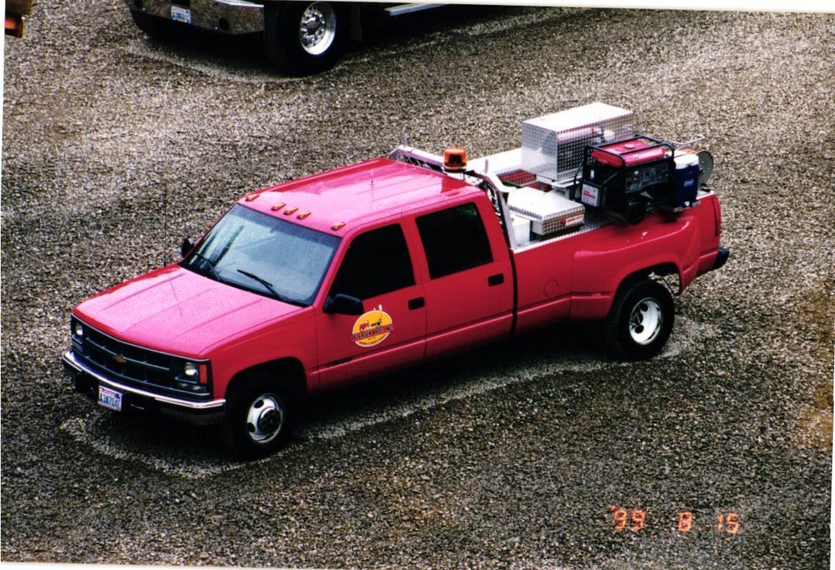 Red Chevrolet dually truck with equipment on the bed, parked on gravel.