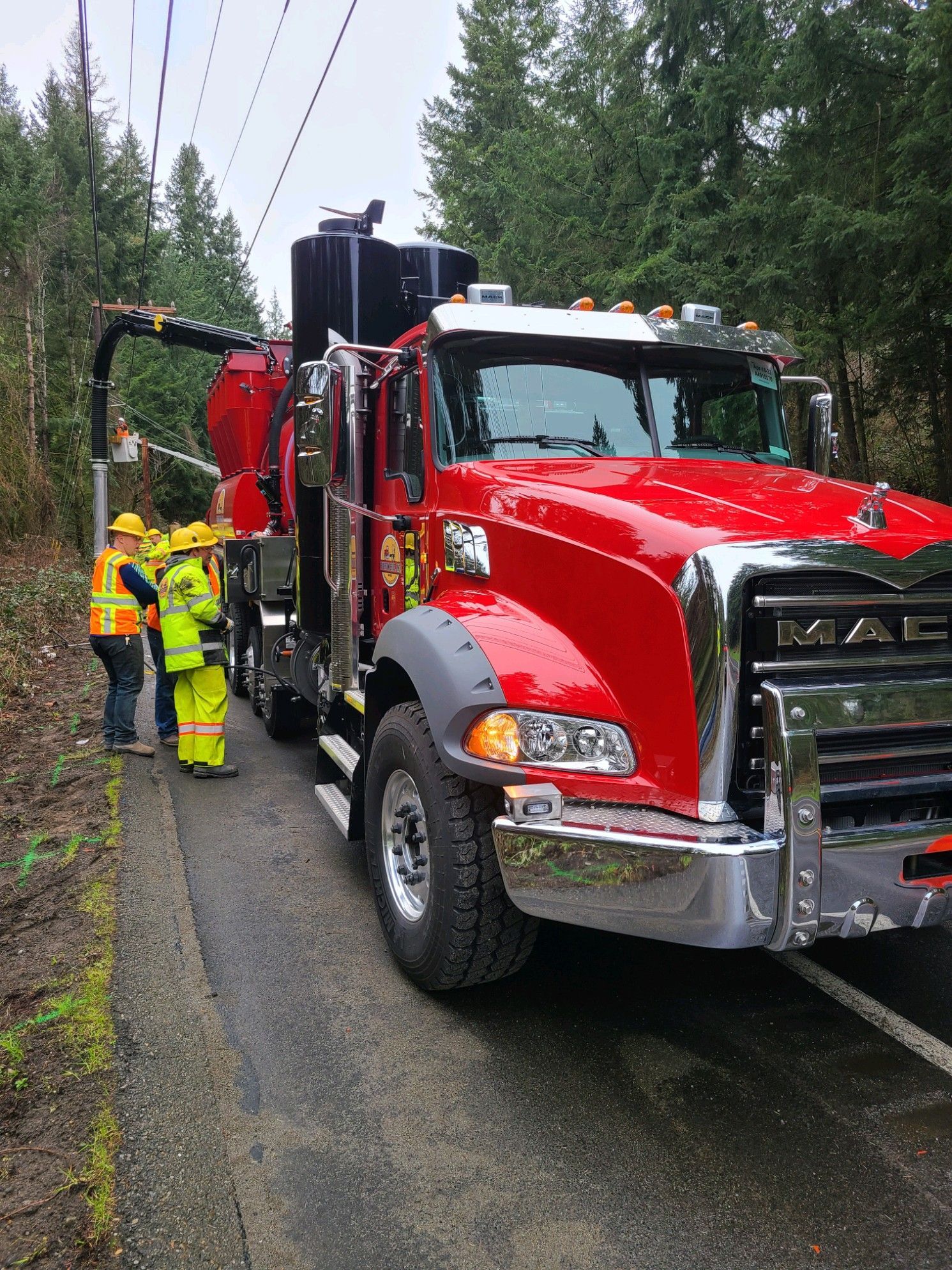 Red Mack truck with workers, vacuum hose near utility pole, on road, surrounded by trees.