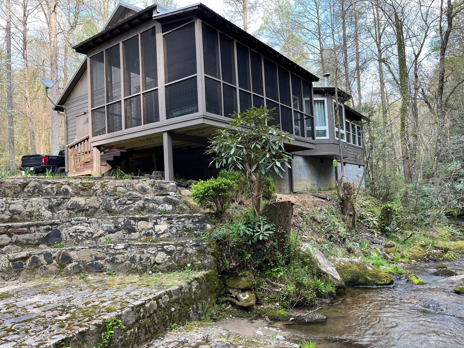 A house with a screened in porch is sitting next to a stream in the woods.
