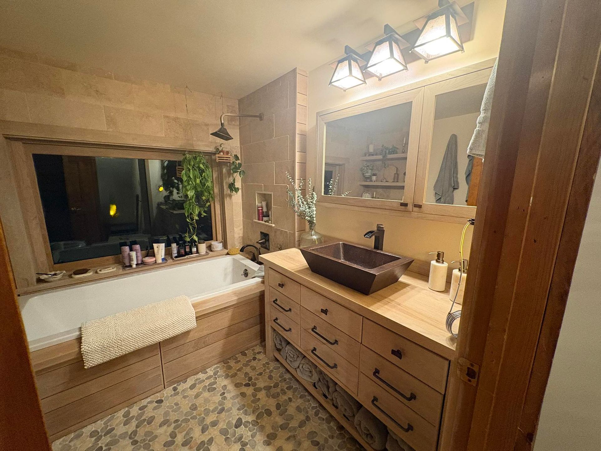 A rustic, wood-toned bathroom featuring a soaking tub, a vessel sink on a wooden vanity, and pebble-tiled flooring.
