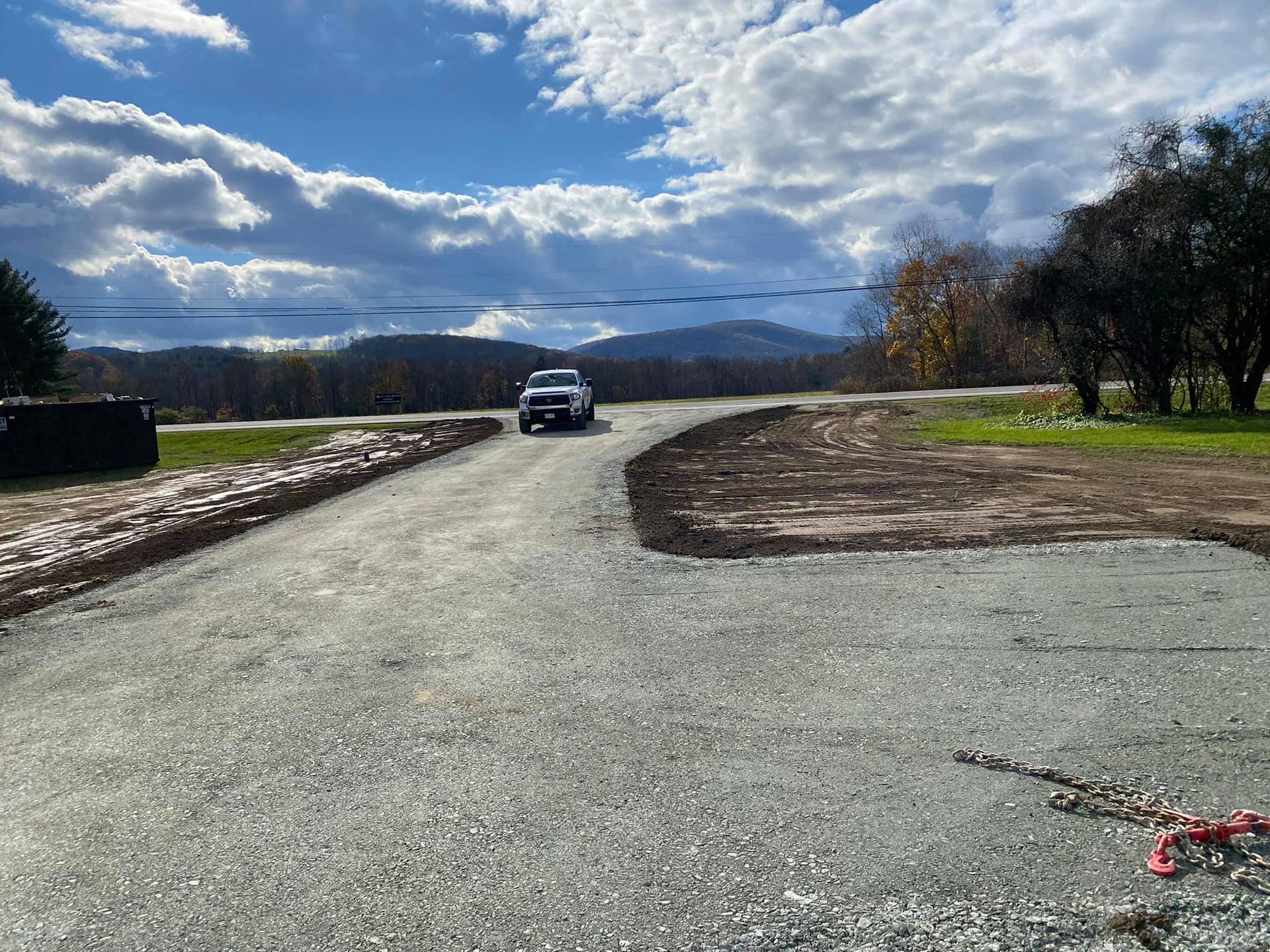 A white pickup truck travels down a newly graveled driveway toward a hillside landscape under a cloudy sky.
