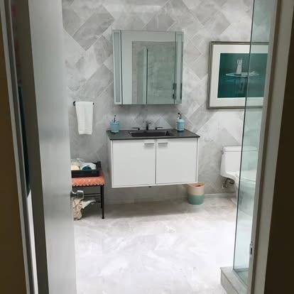 A modern bathroom with a floating vanity, gray tiled walls, a square mirror, and a toilet next to a glass shower door.