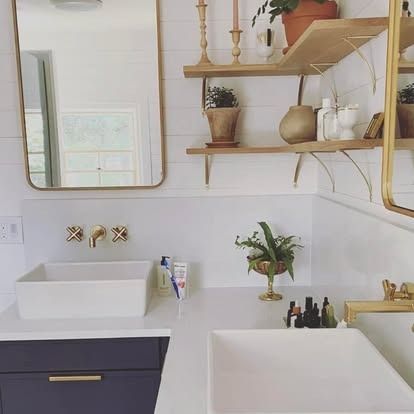 A modern double vanity bathroom with navy blue cabinets, white countertops, brass fixtures, and tiered wooden shelving.