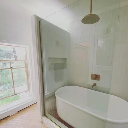 A white freestanding bathtub sits inside a glass shower enclosure with gold hardware in a bright, modern bathroom.