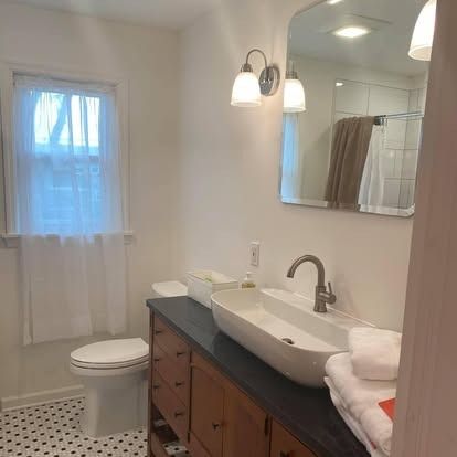 A bright bathroom with a white vessel sink on a wood vanity, a toilet, a window with a sheer curtain, and a wall mirror.