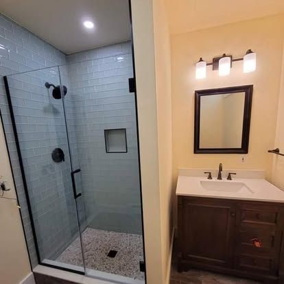 A modern bathroom featuring a walk-in glass shower with blue subway tiles and a wooden vanity with a mirror and lights.
