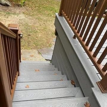 A high-angle view of gray composite deck stairs with brown railings leading down to a patch of grass with fallen leaves.