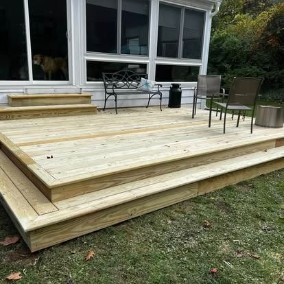 A newly constructed wooden deck featuring steps leading up to a sunroom, with a metal bench and two chairs.