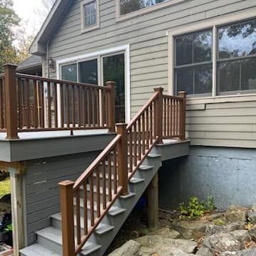 A wooden deck with a staircase and dark brown railing attached to the side of a grey house with a sliding glass door.