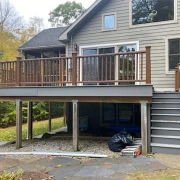 A two-story grey house with a wooden deck, matching railing, and stairs leading down to a stone patio area.