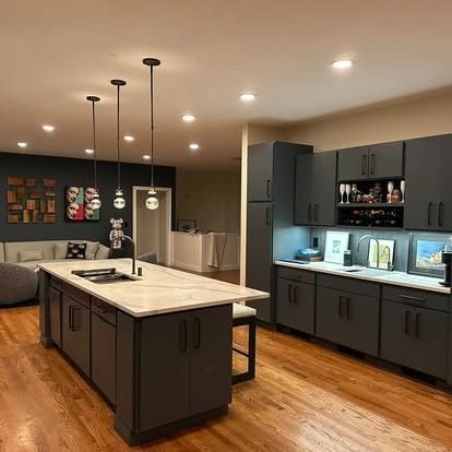 A modern kitchen features dark gray cabinetry, a marble-topped island with pendant lights, and light wood floors.