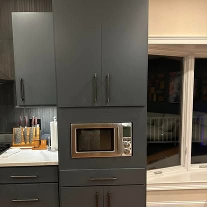 A built-in stainless steel microwave set into matte grey kitchen cabinetry with dark pulls and a nearby knife block.