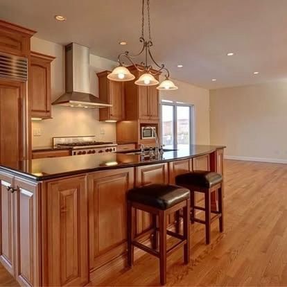 A warm-toned kitchen with wood cabinets, black countertops, a stainless steel range hood, and two bar stools at an island.