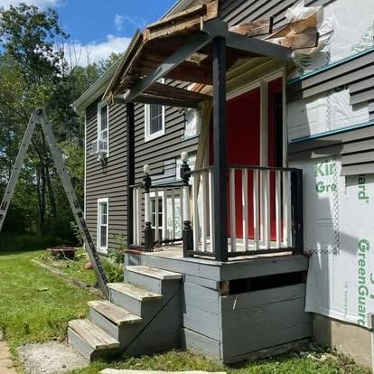 A house entrance under renovation with a red door, gray wooden steps, unfinished siding, and a ladder leaning nearby.
