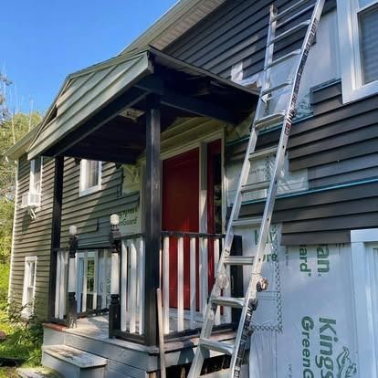 An extension ladder leans against the exterior of a house where dark siding is being installed over house wrap.