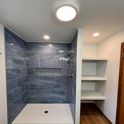 A walk-in shower with gray marbled tiles and a niche, next to built-in white shelves on a light-colored wood floor.