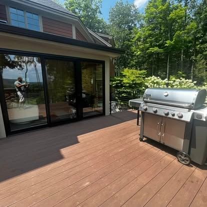 A stainless steel grill sits on a brown composite deck next to a house with large sliding glass doors reflecting the sky.