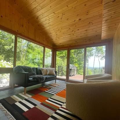Sunroom with wood-paneled walls and ceiling, a dark sofa, two beige armchairs, and a colorful, multi-patterned area rug.
