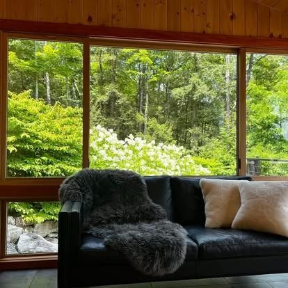 A black leather couch with a grey fur throw and two light cushions, placed in front of a window looking out at trees.