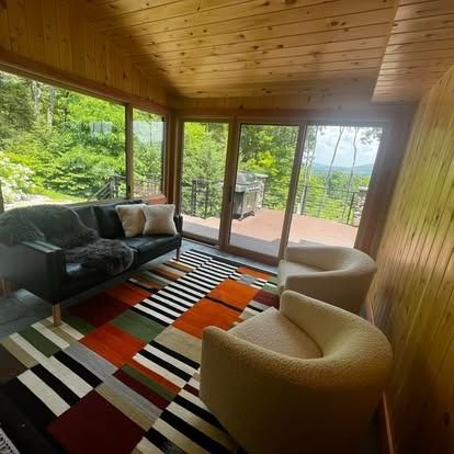 A sunroom with a black sofa, two cream armchairs, and a colorful, geometric rug, overlooking a lush green forest view.
