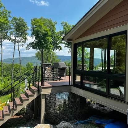 A deck with black railings leads to a house exterior with large, dark-framed windows overlooking a lush mountain view.