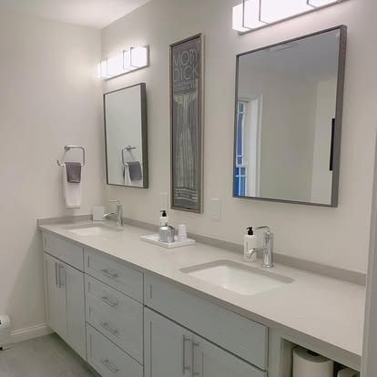 A modern double-sink bathroom vanity with grey cabinets, white countertops, two rectangular mirrors, and wall lighting.