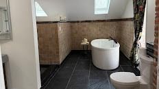 A bright attic bathroom features a white freestanding tub, dark slate-style flooring, tan wall tiles, and a toilet.