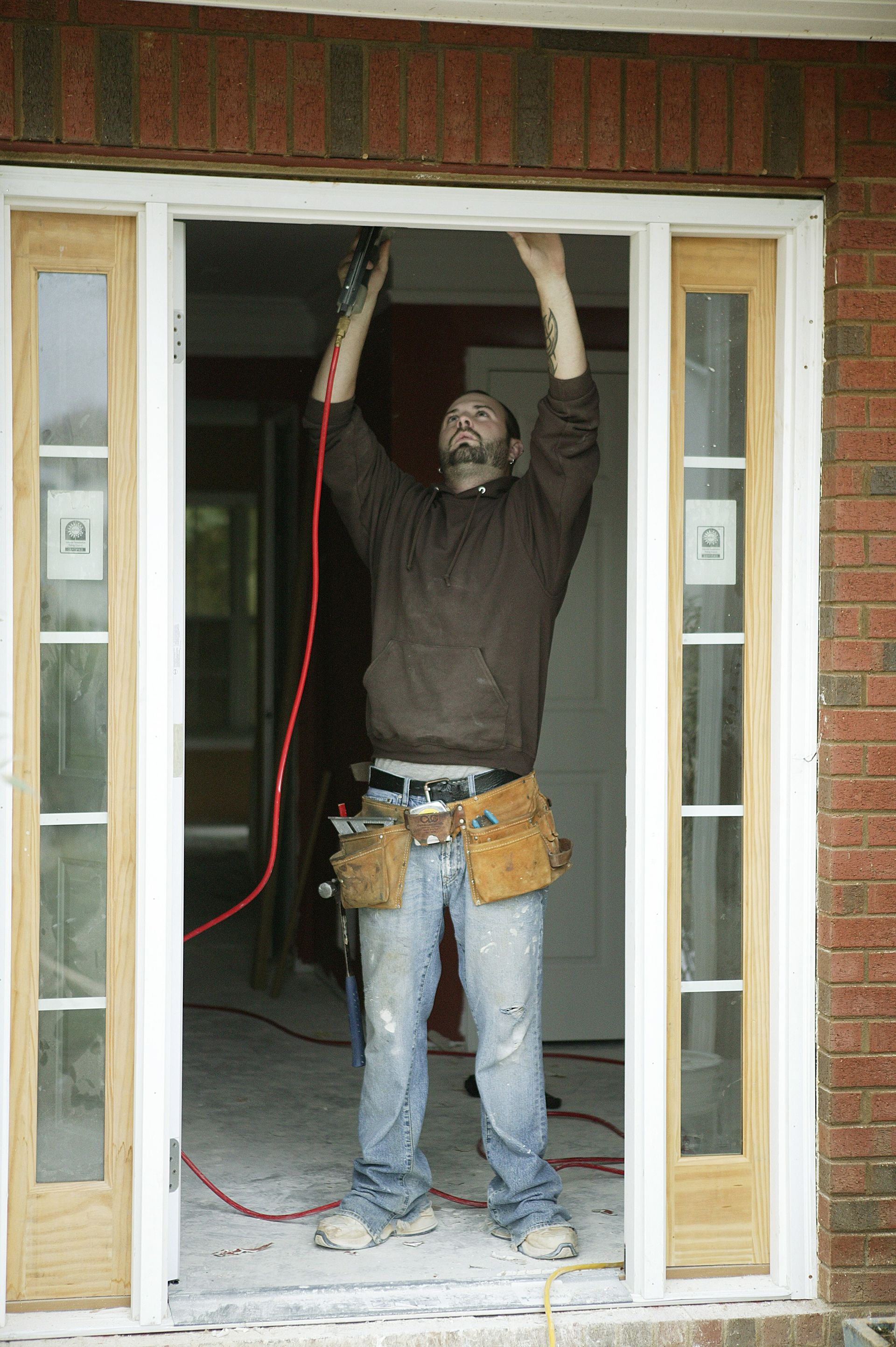 A person in a brown sweatshirt and tool belt installing a new door frame in a brick entryway.