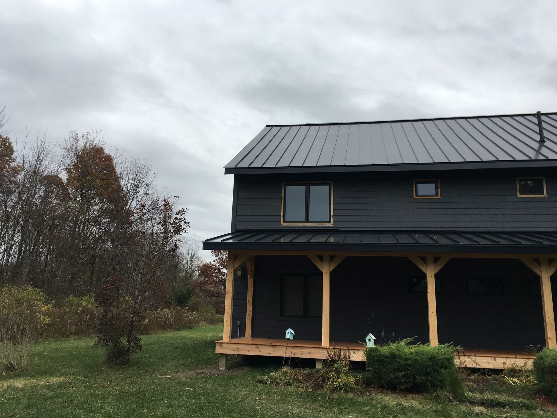 A two-story dark gray house with a black metal roof and a natural wood porch, set against a cloudy sky and autumn trees.