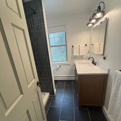 A bright bathroom featuring black tiled floors, a dark walk-in shower, a wood vanity, and three-bulb vanity lighting.