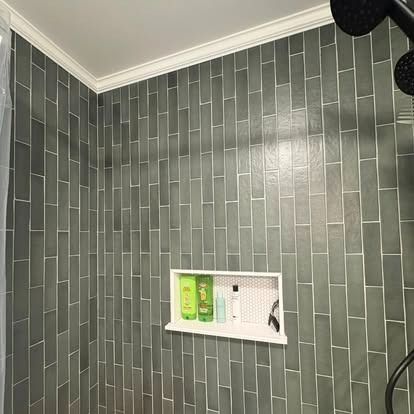 A shower wall featuring vertical, dark sage green subway tiles and a white rectangular recessed shelf with bath products.