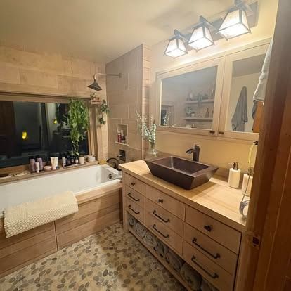 A rustic bathroom with a wooden vanity, pebble-tiled floor, rectangular vessel sink, and a bathtub under a large window.