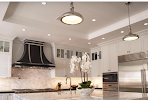 A modern kitchen with white cabinetry, marble countertops, a statement range hood, pendant lighting, and a white orchid.