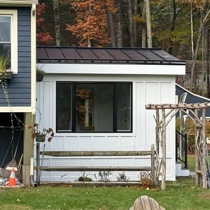 A white, single-story home addition with a dark metal roof, large black-framed window, and rustic wood arbor in a yard.