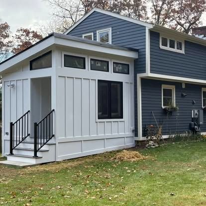 A white modern home addition with black windows and steps attached to a two-story blue house on a grassy lawn.